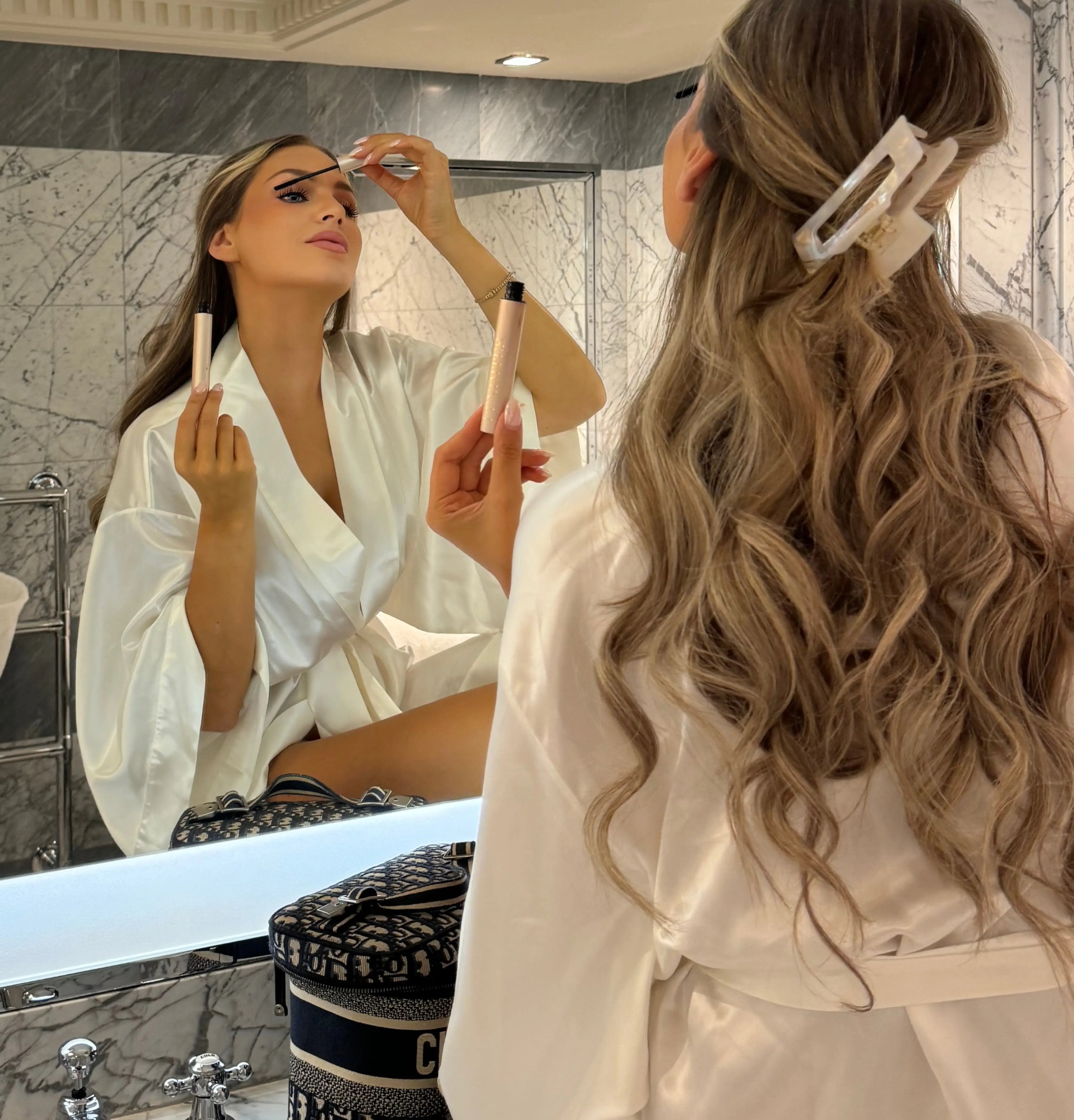 Woman in a white robe applying Lenoites Eyelash Activating Serum Mascara, wearing a Lenoites Eco-friendly Hair Claw, with luxury beauty bags on the counter.